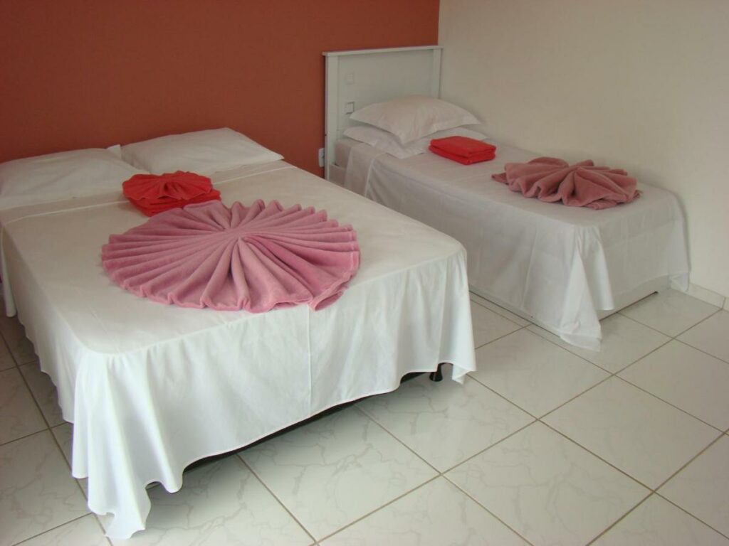 Quarto na Pousada Primavera da Serra com uma cama de casal e uma de solteiro, um terraço, tudo em tons de branco e um rosa claro