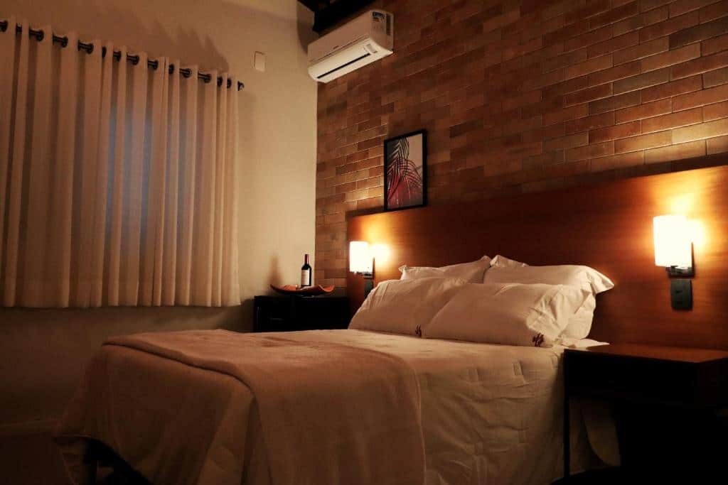 Quarto na Pousada Santa Thereza com uma cama de casal, uma parede de tijolinhos, um ar-condicionado e dois abajures, para representar pousadas em Serra Negra