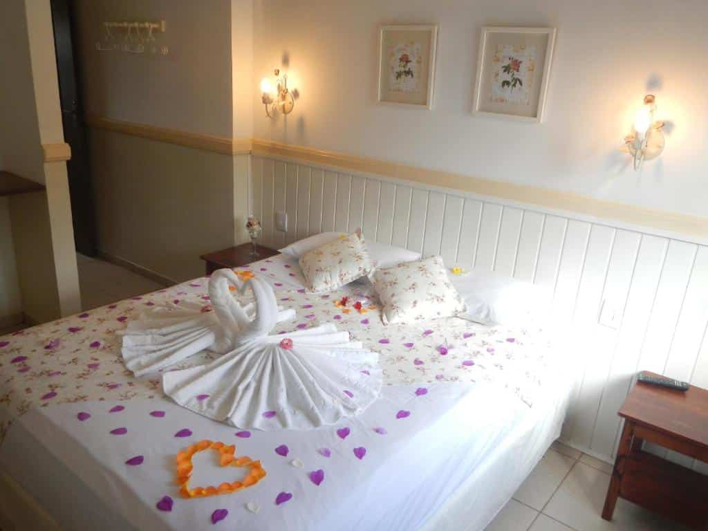 Quarto no Recanto da Praia com uma cama de casal com toalhas e pétalas de rosa, na parede há dois abajures e dois quadrinhos