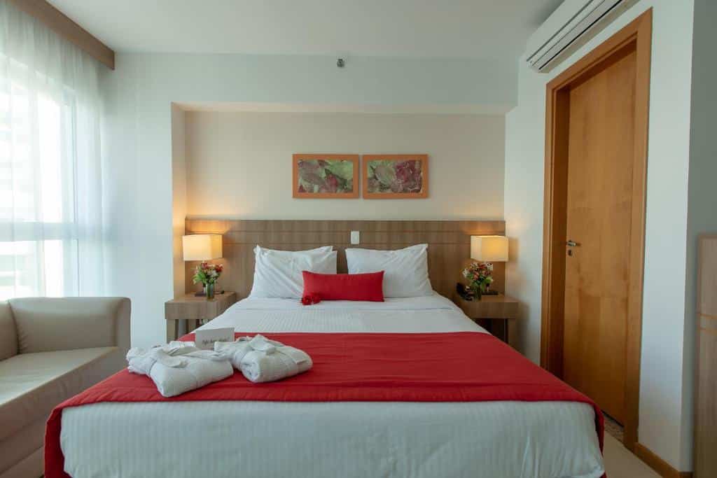 Quarto no Saint Moritz Hplus Express com uma cama de casal, uma cabeceira com abajures embutidos, um ar-condicionado, um sofá, sob a cama há dois roupões, tudo decorado em tons de madeira, vermelho e branco