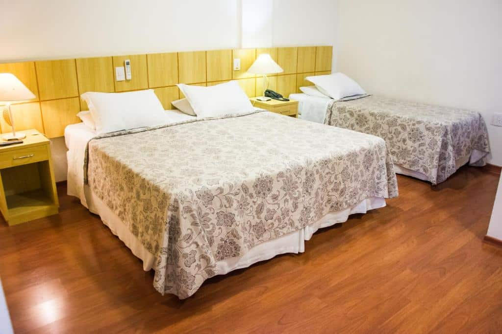Quarto no Serra Negra Palace Hotel com uma cama de casal e uma de solteiro, com chão de madeira, e duas pequenas mesinhas de cabeceira com abajures