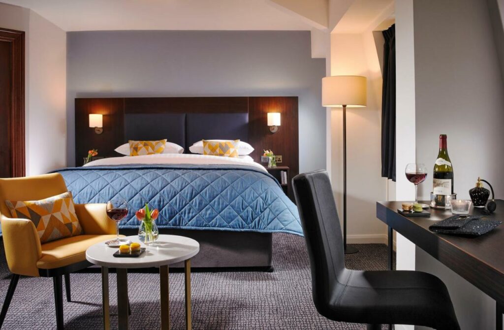 Quarto do Temple Bar Hotel com uma cama de casal, uma pequena mesa de centro com uma poltrona ao lado, uma mesa de escritório com uma cadeira, um abajur de chão e tudo decorado em tons de azul turquesa e amarelo, para representar hotéis em Dublin