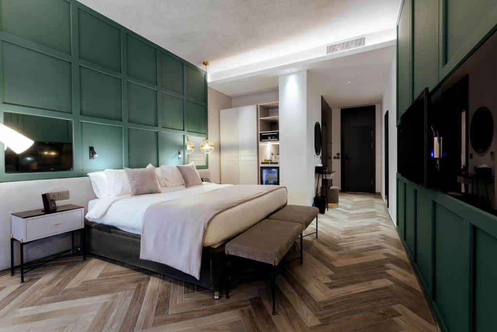 Quarto amplo no The Embassy Valletta Hotel, piso de madeira em vários tons, uma cama de casal, uma televisão, um armário, frigobar, tudo pintado em tons de verde musgo e os móveis brancos, para representar hotéis em Malta