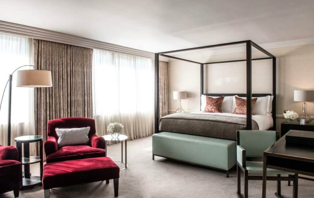 Quarto no The Westbury Hotel com uma cama de casal, duas janelas amplas, uma poltrona, uma luminária de chão, uma mesa de escritório e uma cadeira, tudo em tons de azul bem claro, cinza e branco