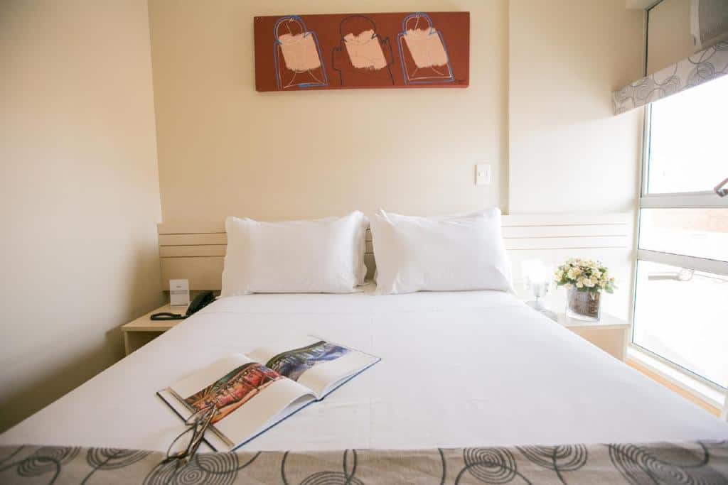 Quarto do Verona Hplus Long Stay com uma cama de casal, uma cabeceira com duas mesinhas, um vaso de flor, uma janela, um quadro e as roupas de cama são brancas, para representar hotéis em Brasília