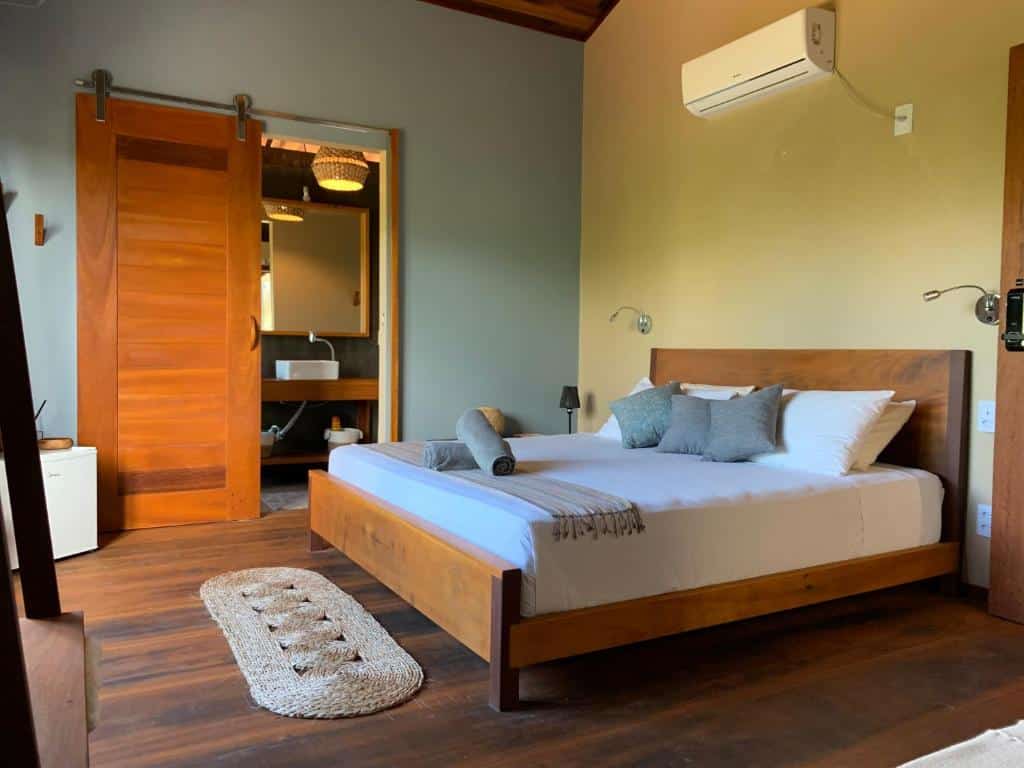 Um quarto da Vila das Águas com uma cama de madeira, roupa de cama cinza, uma porta deslizante e um ar-condicionado na parede.