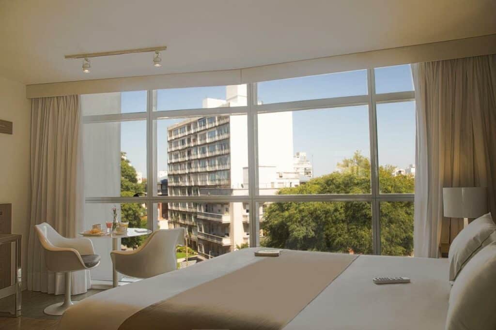 Quarto do Vivaldi Hotel Loft Punta Carretas com uma janela que vai do teto até o chão dando vista pra cidade, uma cama de casal, uma pequena mesa com duas cadeiras e um abajur, para representar hotéis em Montevidéu