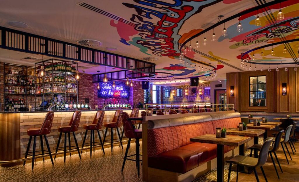 Espaço para refeições no Hard Rock Hotel Dublin com teto desenhado e colorido, um bar iluminado, cadeiras vermelhas e mesinhas de madeira