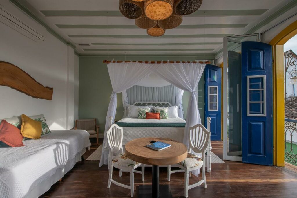 Qaurto do Sandi Hotel com duas sacadas, uma cama de casal, uma mesinha com duas cadeiras, um sofá, tudo decorado em tons de branco e azul, o chão é de madeira