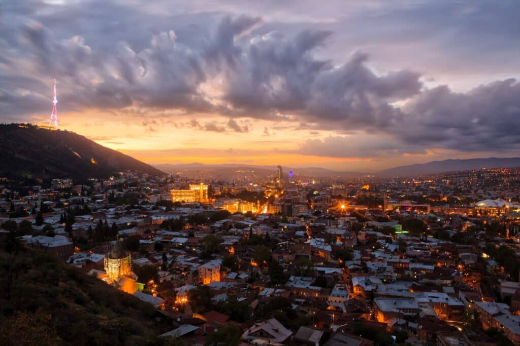 Vista de cima de Tbilisi, capital da Geórgia, para ilustrar o post de seguro viagem Geórgia. É final de tarde e o sol está se pondo. - Foto: Jaanus Jagomägi via Unsplash