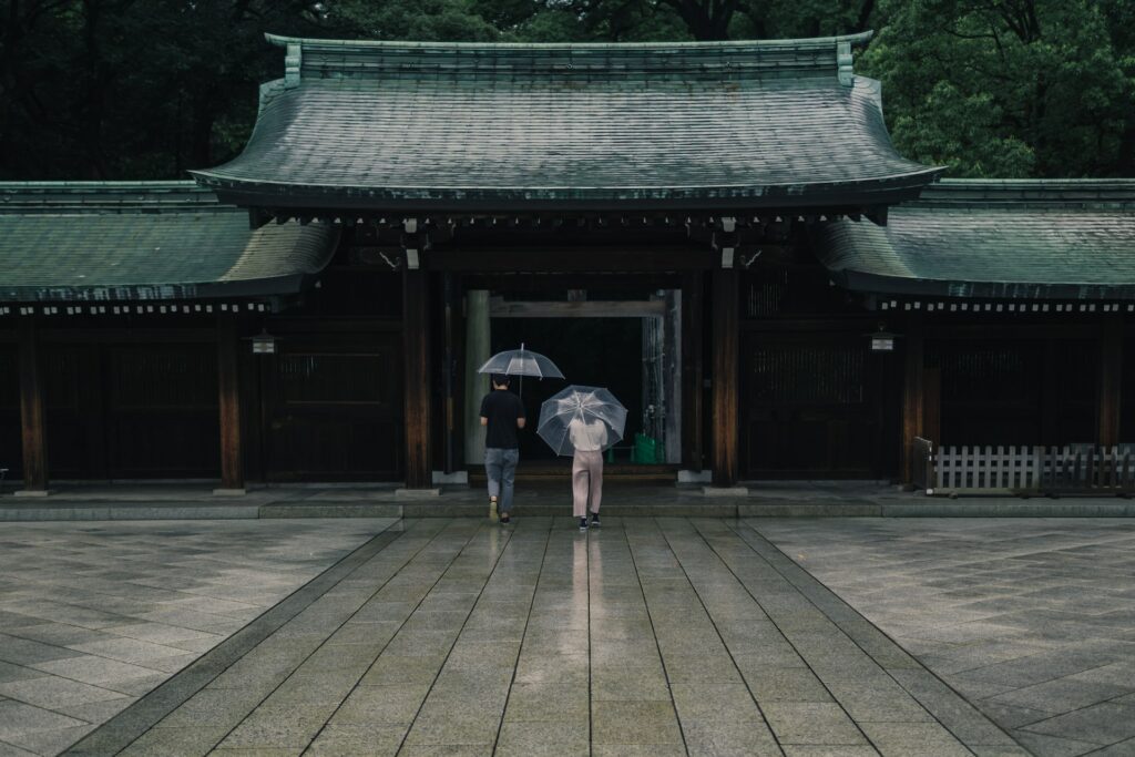 Duas pessoas com guarda-chuva entrando no templo de Menji Jingu, em dia de céu nublado e com chuva fina caindo, para representar o seguro viagem Tóquio.