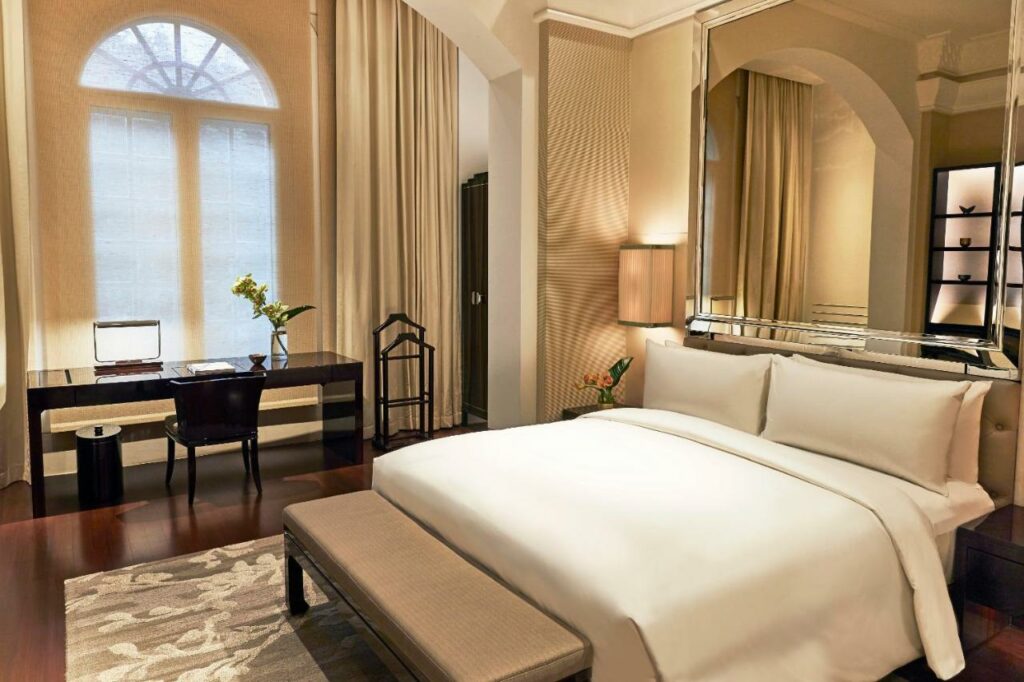 Quarto de hotel em Singapura com cama de casal com edredom branco, tapete, escrivaninha com computador, espelho e uma janela com cortina.