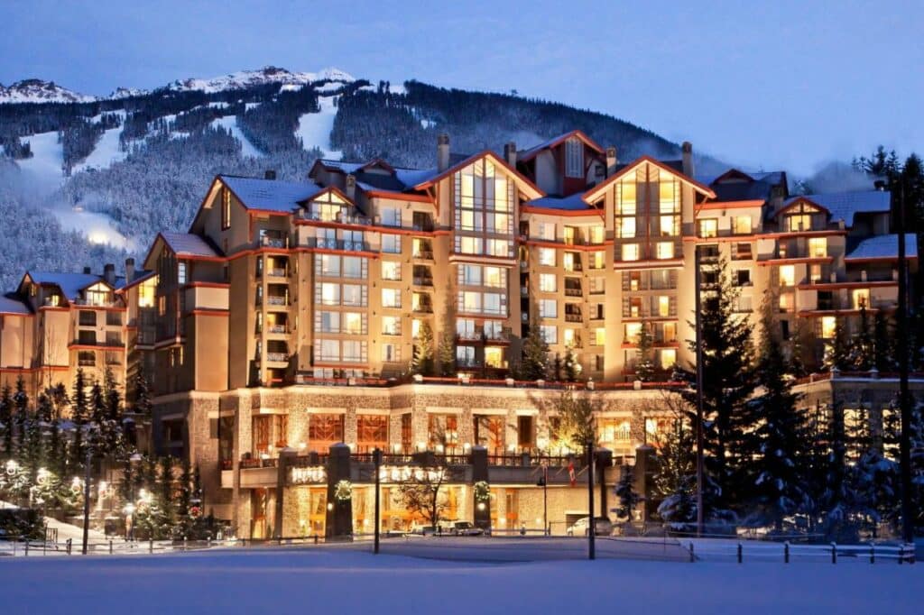 Fachada do hotel The Westin Resort & Spa em Whistler. Iluminado a noite cercado por vegetação e neve com uma montanha com neve e vegetação ao fundo.