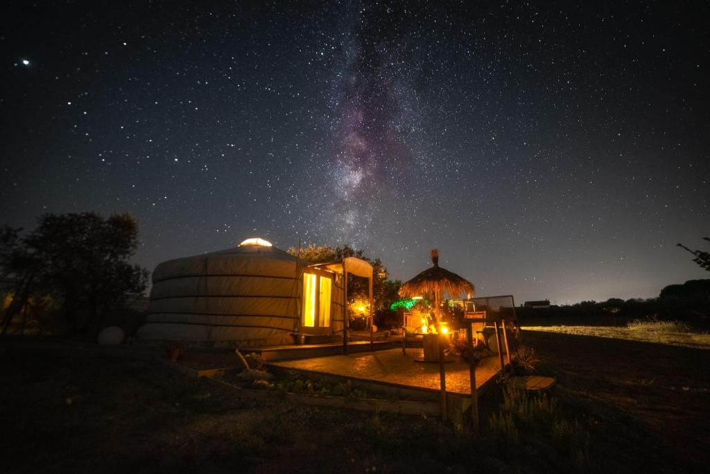 Camping com fogueira acesa, em uma noite estrelada, sendo a barraca redonda e espaçosa com luzes ligadas dentro, gramado baixo em volta, com algumas árvores