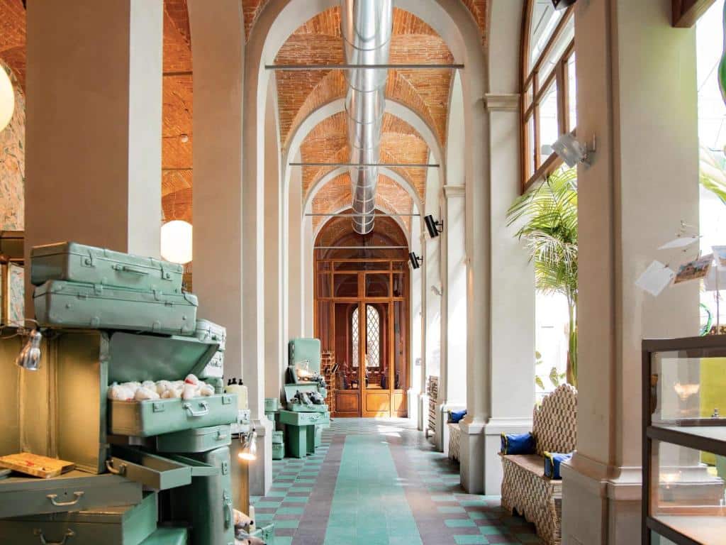 Corredor do 25hours Hotel, em Florença, com malas verde água decorando o espaço, além de bancos entre colunas altas e uma porta de madeira grande no fim do corredor