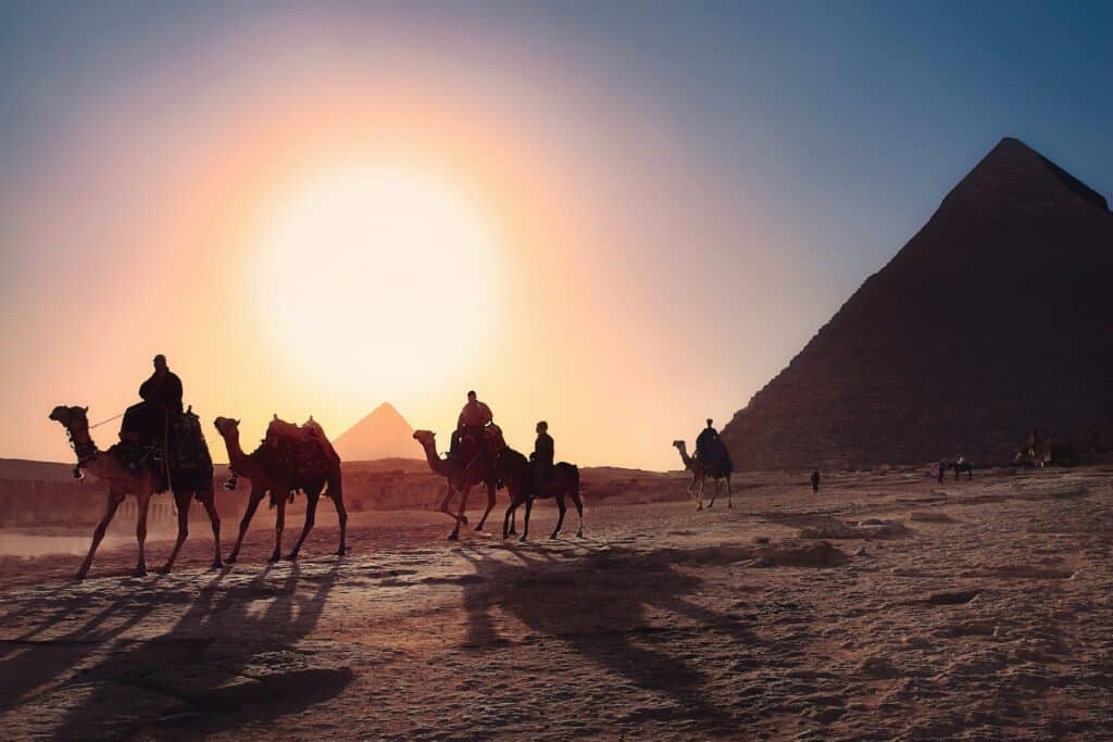 pessoas andando de camelo ao pôr do sol no deserto com as Pirâmides de Gizé ao fundo, no Egito