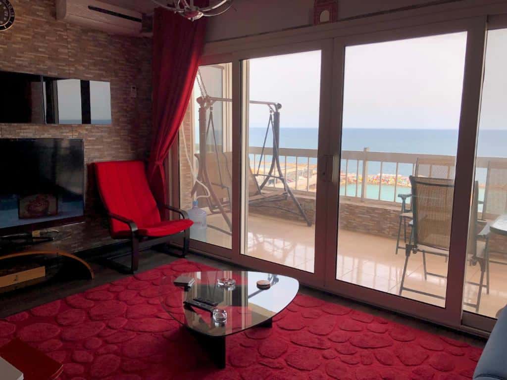 vista do apartamento Blue Bay Beach-Families only, em Alexandria, todo decorado em vermelho com poltrona, mesa de vidro e vista do rio da sacada, com balanço e cadeiras