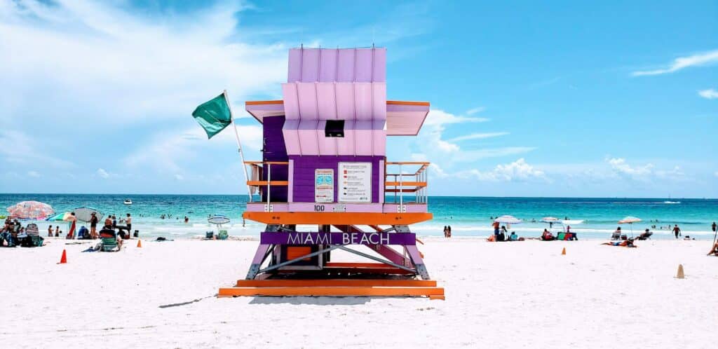 vista da Miami Beach com água azul e areia branca com uma barraquinha em tons de rosa e roxo bem bonitinha em madeira que pode ser aproveitada com o chip celular Miami