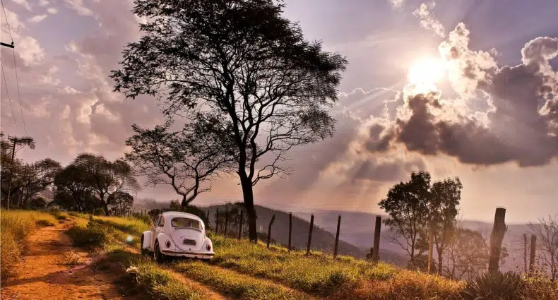 Vista de um Fusca branco em estrada de terra com uma árvore grande e vista do Pico do Olho d'Água com o sol em tons de roxo em Mairiporã
