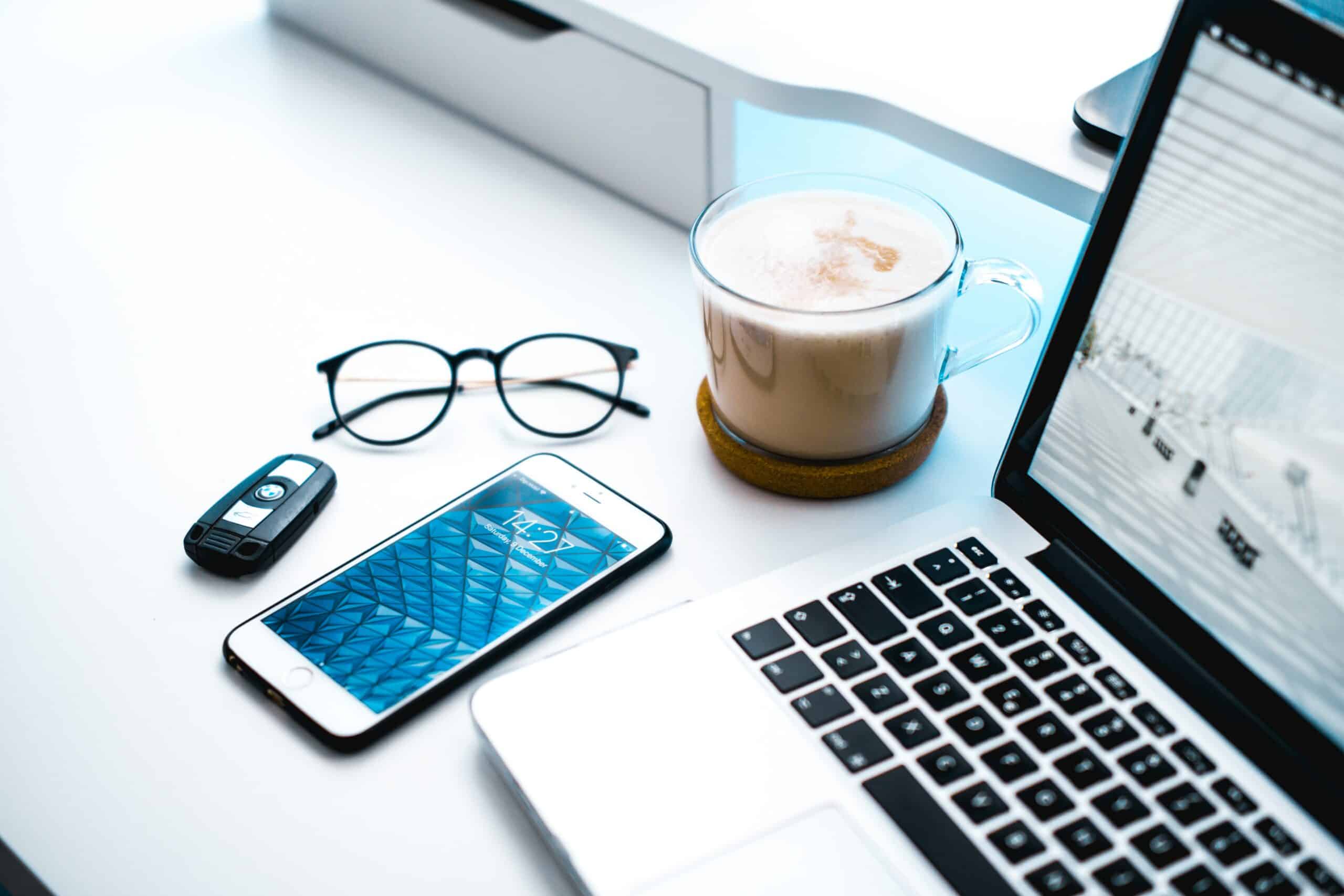mesa de trabalho branca com um óculos feminino, um chave de carro, um celular iphone, uma xícara trabsparente com capuccino e um computador