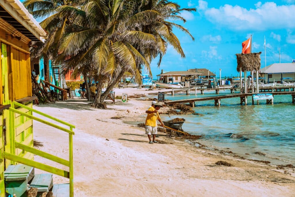 Homem de camiseta amarela, bermuda branca e chapéu bege limpando a areia de uma praia para ilustrar o post seguro viagem Belize. Ao lado esquerdo há uma casa amarela e árvores, e do direito uma ponte de madeira acima do mar cristalino. - Foto: Meritt Thomas via Unsplash