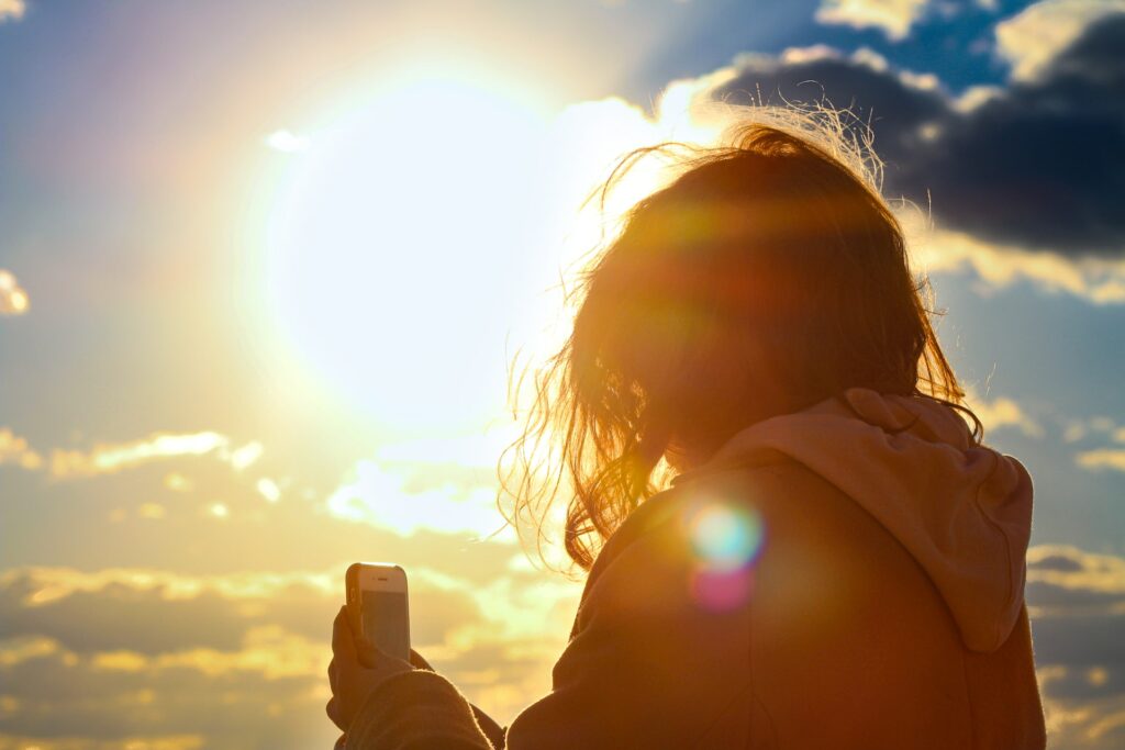 Uma mulher de costas usando um moletom com capuz segurando um celular com o sol batendo bem no rosto dela