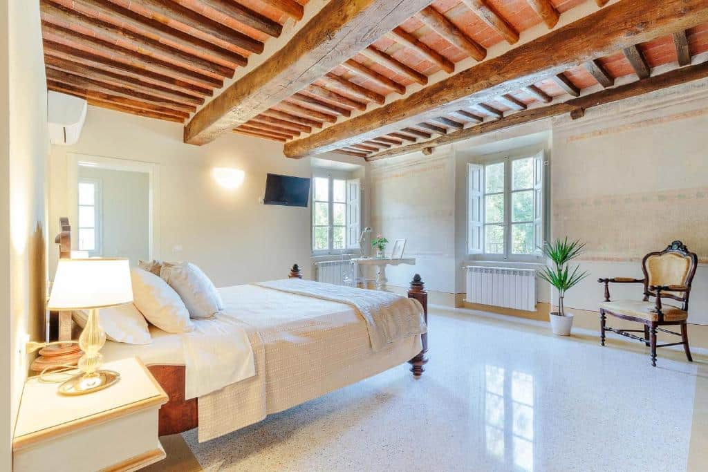 Quarto de um dos hotéis na Toscana, o Antica Pergola di San Giusto, com teto de madeira, duas janelas, poltrona de época, pequena mesa redonda com vaso de flor em cima, cama de casal e mesinha ao lado com abajur em cima