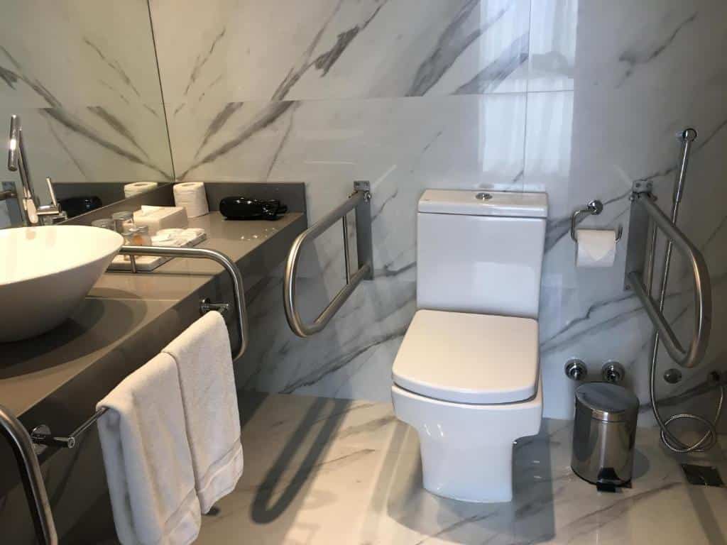 Banheiro do AQ Tailored Suites com barras de apoio do lado do vaso sanitário, uma pia mais baixa com barras de apoio também, com toalhas penduradas, para representar os hotéis em Buenos Aires