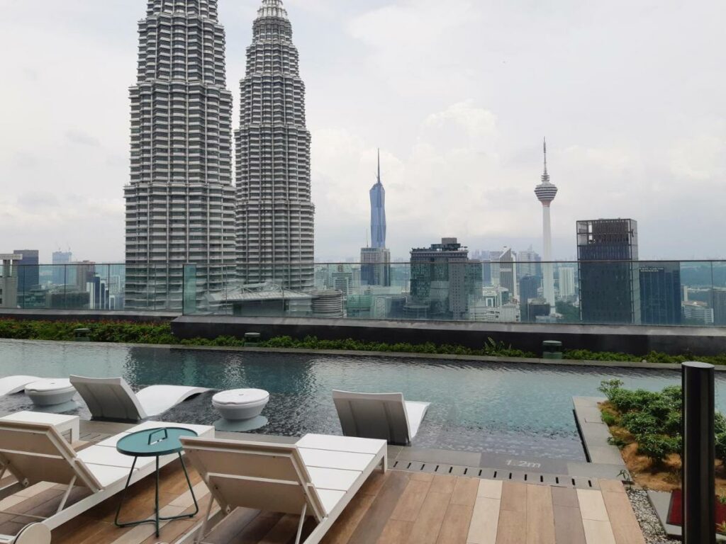 Área da piscina do hotel Star Luxury Suites KLCC em Kuala Lumpur, cadeiras em frente a uma piscina com algumas vegetações ao em torno e vista para prédios e arranha céus modernos da cidade.