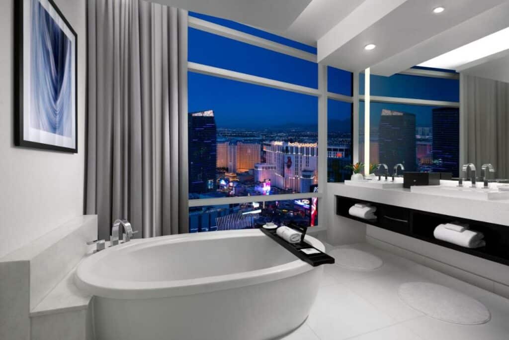 Um banheiro amplo no ARIA Resort & Casino com uma janela panorâmica com vista para os casinos, com uma banheira oval e do lado direito tem duas pias e um móvel com toalhas disponíveis, o piso é branco