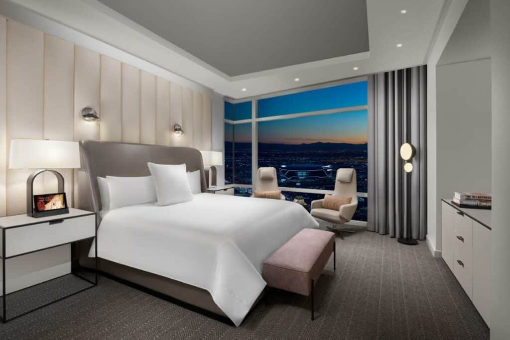 Quarto no ARIA Resort & Casino com uma janela panorâmica com cortinas cinzas, duas poltronas modernas beges, uma cama de casal, duas mesinhas de cabeceira com uma gaveta, o carpete cinza e um móvel do lado direito, para representar resorts em Las Vegas