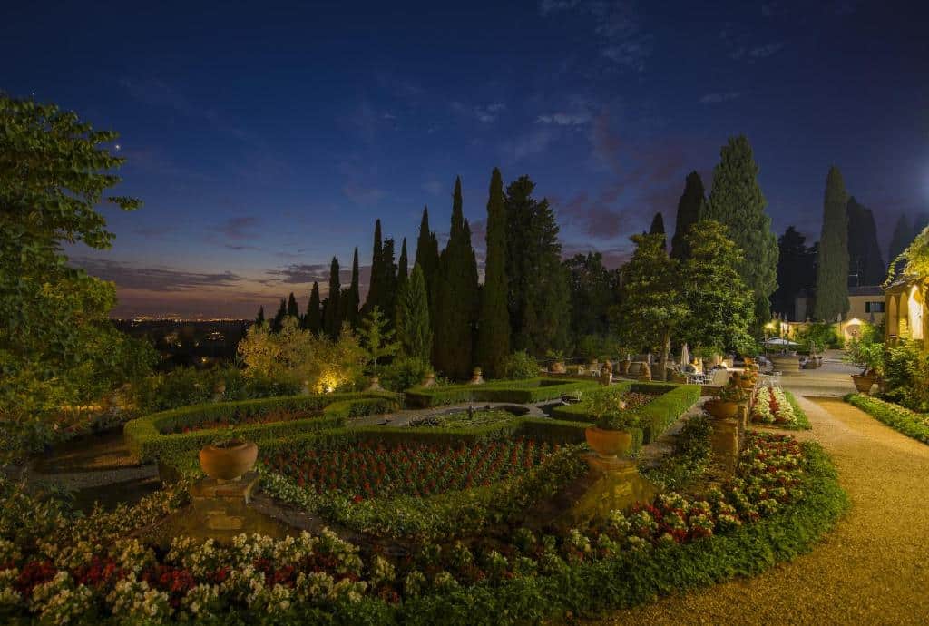 Jardim do Art Hotel Villa Agape, num começo de noite, com céu azul e roxo, e bastante verde, incluindo árovres altas, flores vermelhas e gramado verde