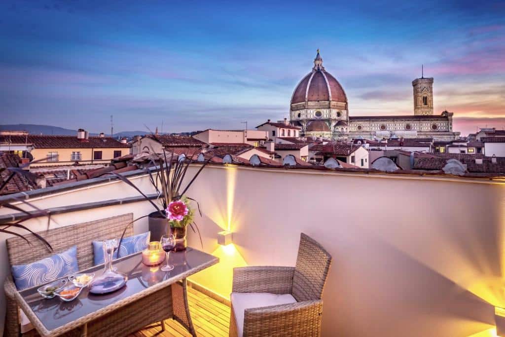 Varanda do Arte' Boutique Hotel, em Florença, com uma mesa pequena retangular com taça de vinho, vaso de flor e petiscos em cima, duas cadeiras, e vista para o telhado das casas da região, além da cúpula de uma igreja, e o céu num começo de noite na tonalidade azul com rosa