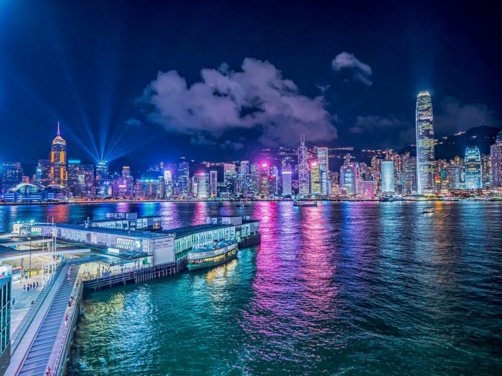 Baía de Victoria, ao anoitecer com céu iluminado por luzes coloridas cercado por construções de prédios modernos para representar o seguro viagem Hong Kong.