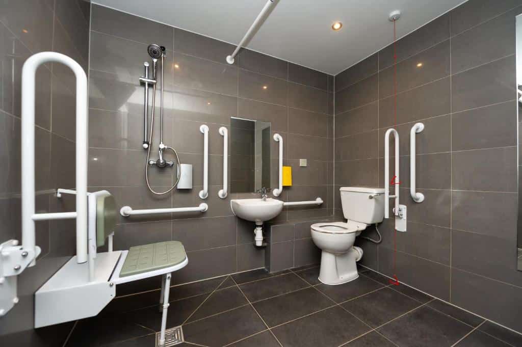 Banheiro adaptado do Staycity Aparthotels, com paredes e chão num tom cinza escuro, além de vaso, pia, cadeira de banhro e chuveiro com barras de apoio ao lado e bastante espaço entre cada objeto