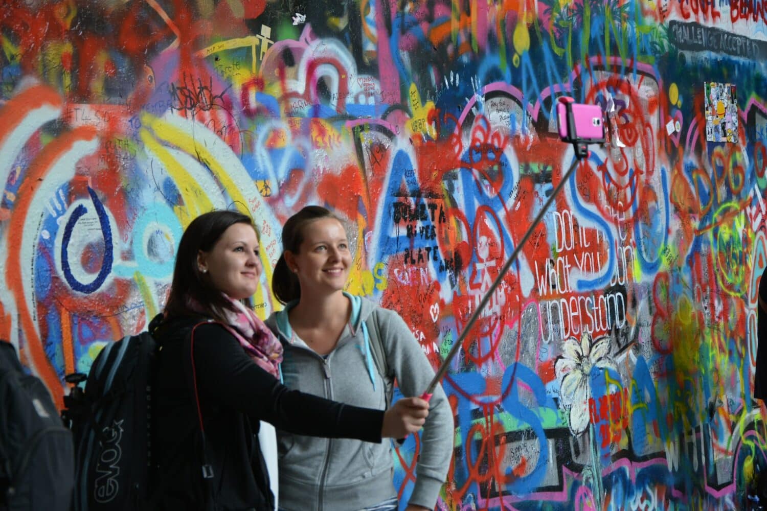 Duas moças, uma loira e de moletom cinza e outra de cabelo castanho e blusa preta, tirando uma selfie em frente um muro todo colorido em Praga