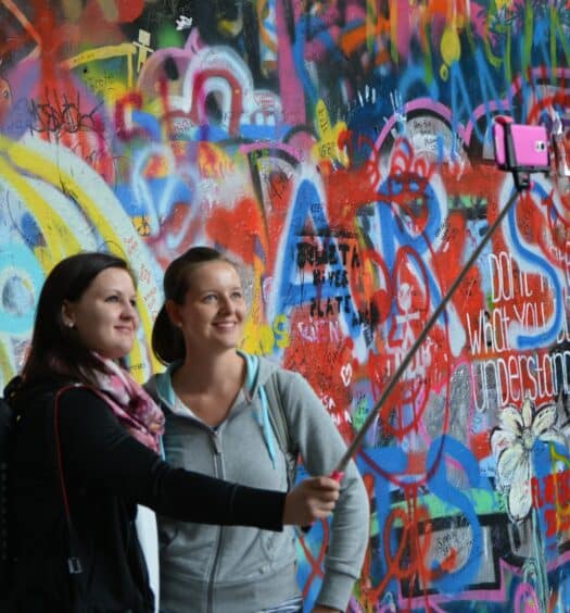 Duas moças, uma loira e de moletom cinza e outra de cabelo castanho e blusa preta, tirando uma selfie em frente um muro todo colorido em Praga