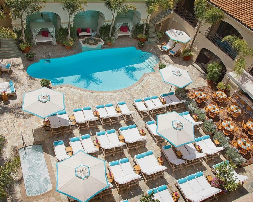 Vista aérea da piscina do Beverly Wilshire, A Four Seasons Hotel em tamanho médio, ao redor, é possível ver espreguiçadeiras brancas com almofadas azuis claras, além dos guarda-sóis e muitas árvores ao redor da piscina