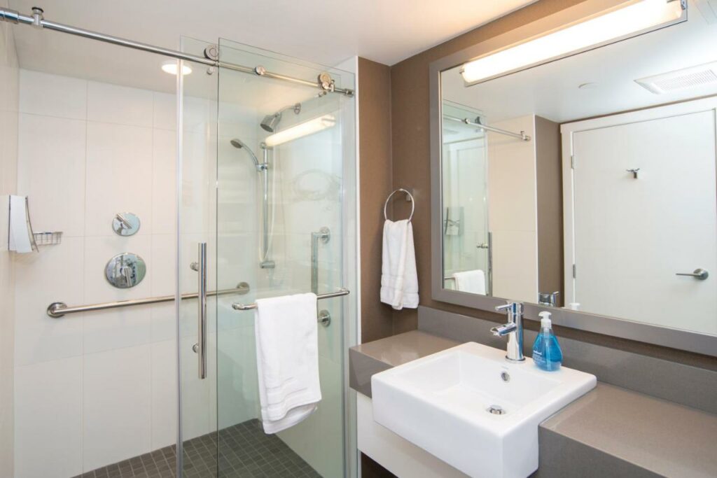 Banheiro no Blue Horizon Hotel com um box cheio de barras de apoio, do lado esquerdo, há uma pequena pia e um espelho sob ela