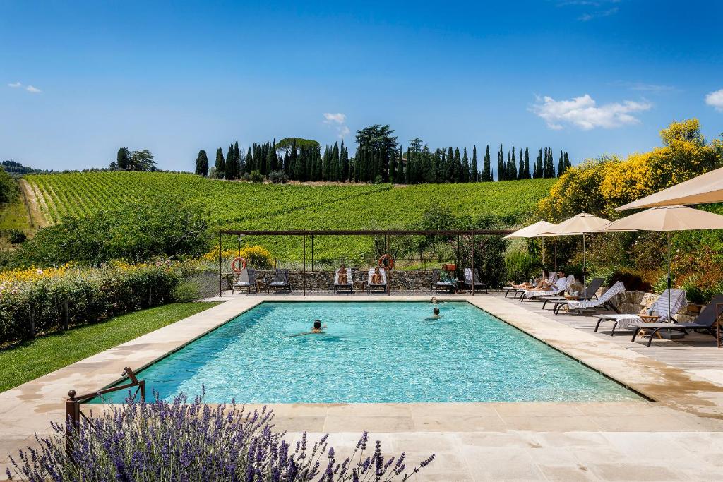 Piscina retangular grande de um dos hotéis na Toscana, o Borgo Del Cabreo, com espreguiçadeiras em volta e vista do campo amplo verde do local, com plantações de uva, árvores e flores lilás e amarelas