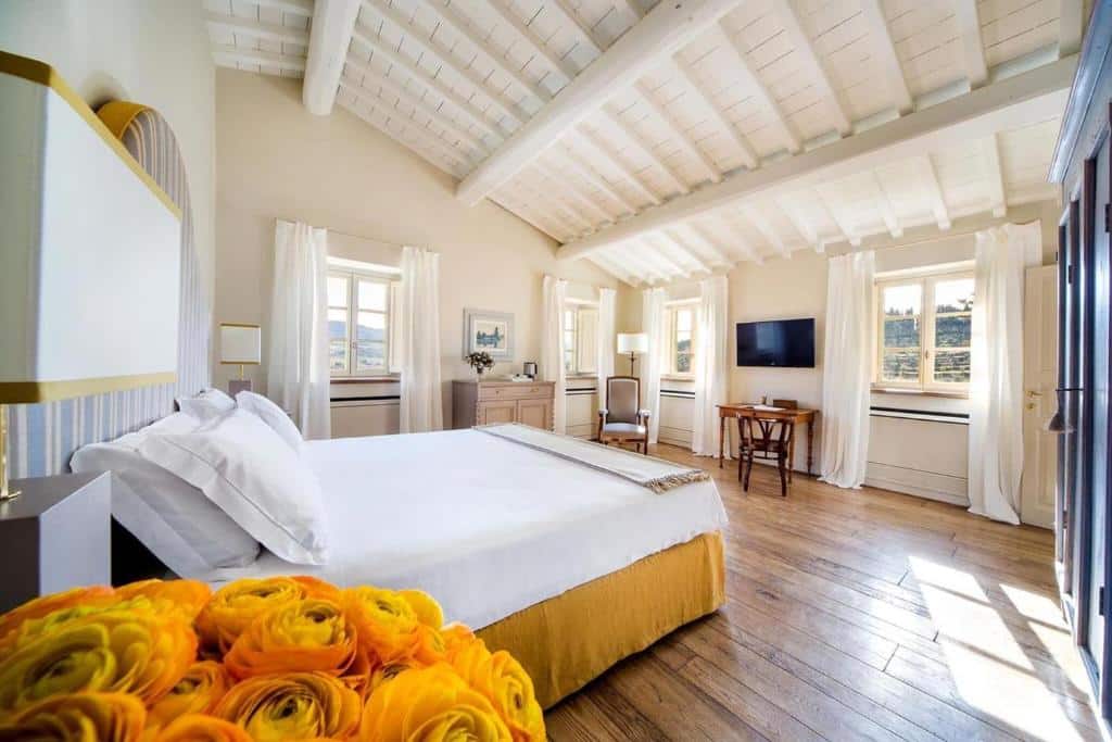 Quarto duplo deluxe do Borgo Del Cabreo, com 30 m², cama de casal grande, flor amarela, TV, mesinhas retro, poltrona e uma cadeira. Há bastante espaço entre a cama e as paredes