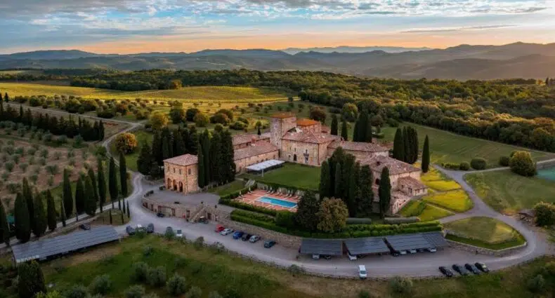 Vista aérea do hotel cinco estrelas Borgo Scopeto, com bastante vegetação em volta, céu apontando um fim de tarde com pouco sol e tonalidade arrasada com azul, e prédios da propriedade com aspecto de castelo medieval, para representar hotéis na Toscana.
