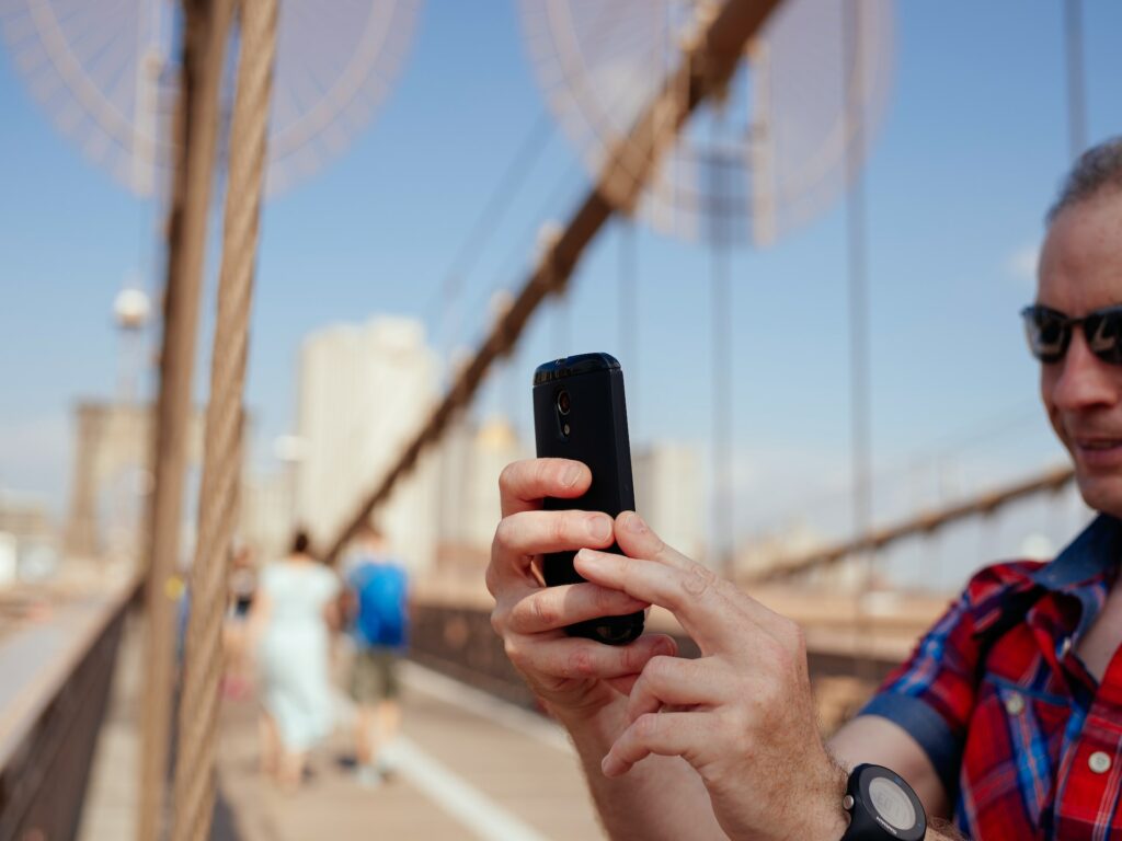 Homem na Ponte de Brooklyn, em Nova York, segurando um celular com as mãos, com um relógio no pulso, olhando para a tela e sorrindo