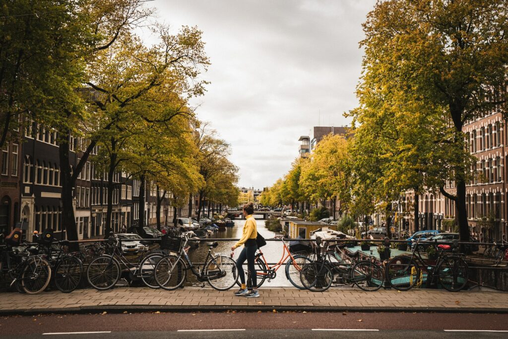 Mulher de blusa amarela encostando uma bicicleta em meio a diversas outras para ilustrar o post chip de celular para Holanda. Há barcos no canal e árvores em sua margem. - Foto: Liam McGarry via Unsplash