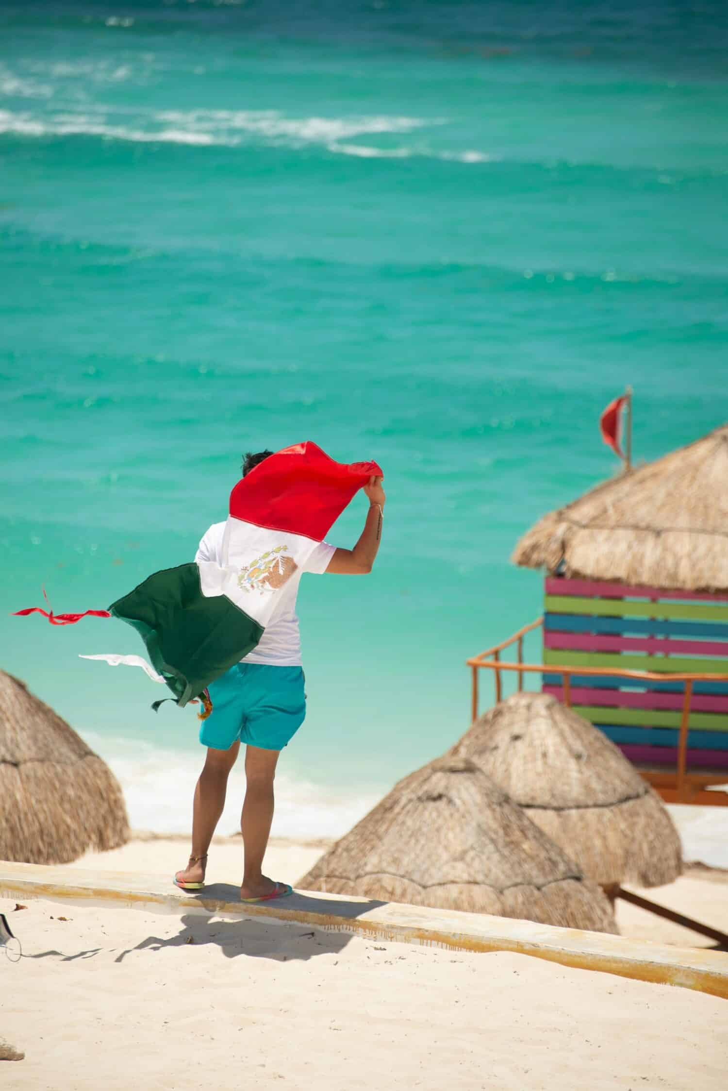 pessoa de costas segurando a bandeira do México (três listras - verde, branco e vermelha, o branco tem um desenho) em frente a praia e alguns quiosques na praia em Cancun