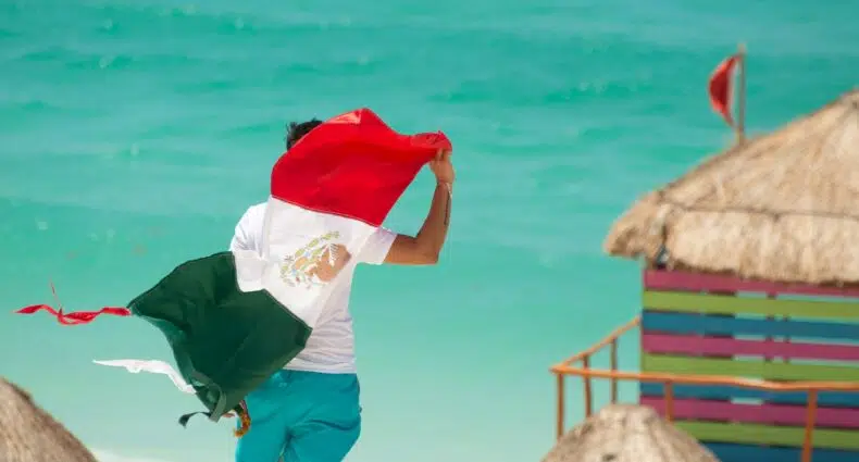 pessoa de costas segurando a bandeira do México (três listras - verde, branco e vermelha, o branco tem um desenho) em frente a praia e alguns quiosques na praia em Cancun