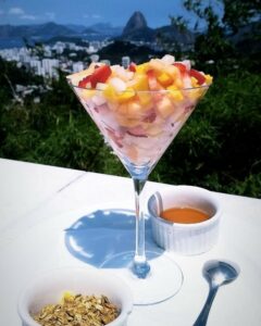 Salada de frutas na taça da Casa Marques, com um pote de mel e um de granola ao lado. Pode-se ver o Pão de Açúcar ao fundo da foto.