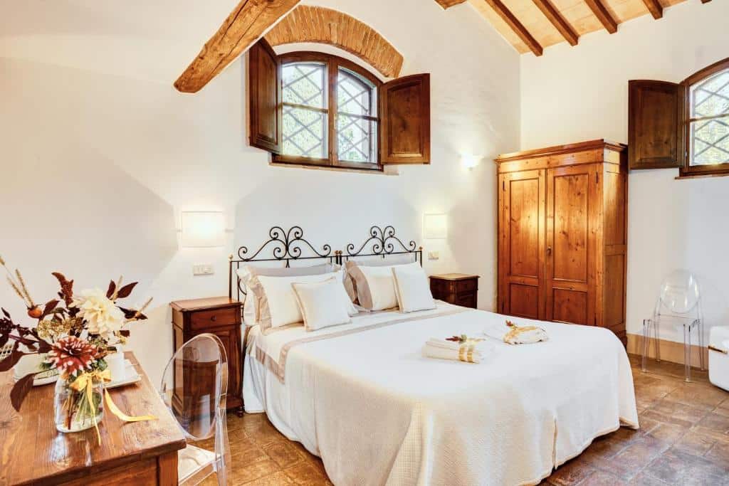Quarto do Castello di Spedaletto da Laura, com 18 m², com cama de casal com travesseiros, almofadas e toalhas enroladas em cima, mesa de quadrada de madeira com vaso de flor em cima e cadeiras transparentes