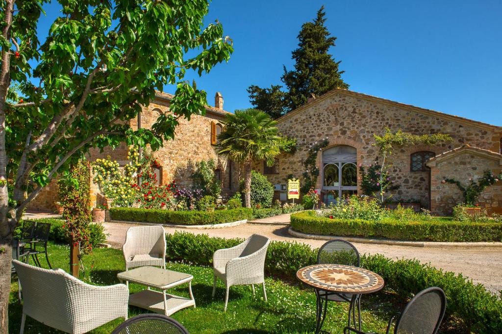 Área externa do Castello di Spedaletto da Laura, um dos hotéis na Toscana, com diversos jardins floridos, grama verde, cadeiras espalhadas no meio da natureza, céu azul e entrada do prédio da propriedade