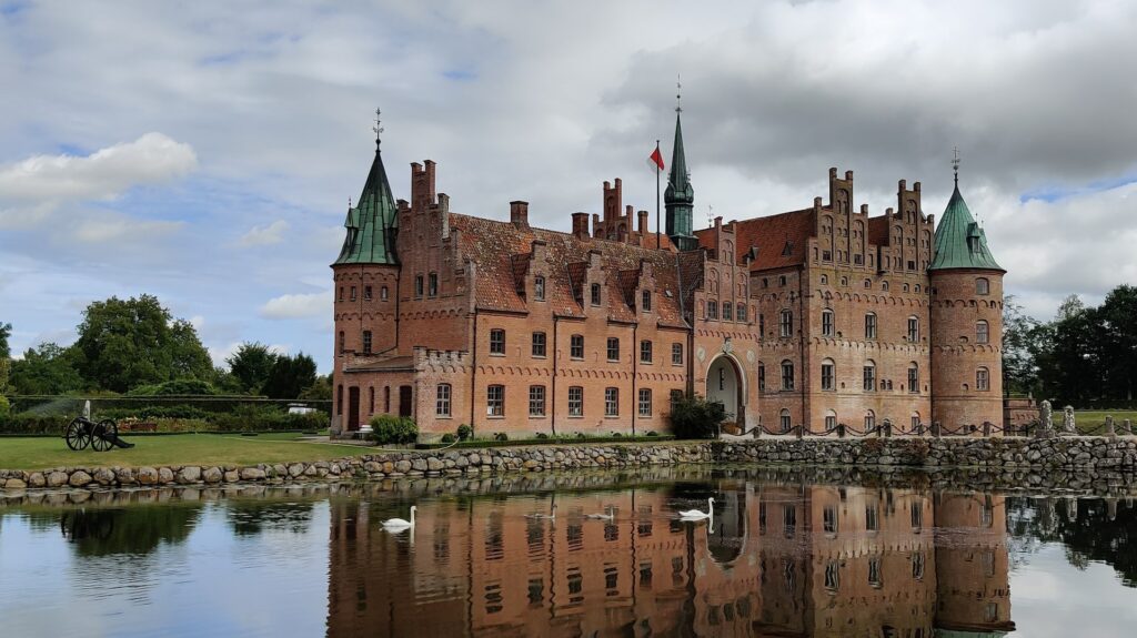 Castelo de cor avermelhada na frente de um lago com gansos para ilustrar o post chip de celular para Dinamarca. Ao fundo há grama e o céu está nublado. - Foto: Andreas Helveg Rudolph via Pexels
